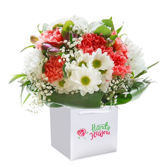 Within a crisp, square white gift bag, a lavish bouquet blooms-a testament to the refined artistry and heart of Yiewsley Florist. The arrangement's centre bursts with full, frilled carnations in coral, peach, and deep crimson, each petal subtly layered to evoke an abundant, textured tapestry. White daisies with luminous chartreuse centers bring a lively contrast and youthful air, reminiscent of sunlit afternoons around Yiewsley's parklands. Wispy clouds of white baby's breath infuse the bouquet with a gentle softness, gracefully connecting each vibrant flower. Here and there, tight purple-tinted buds emerge, inviting a quiet fascination and symbolizing the potential for new beginnings in the ever-changing community landscape. The whole arrangement is embraced by broad, shiny green leaves and structured foliage, framing the blooms and drawing the eye to every tonal shift. Seated in the pristine gift bag, finished with strong ribbon handles, the flowers are illuminated by natural light against a pure white setting-emphasizing their freshness and contemporary grace. Specially designed for prompt and personal delivery across the Yiewsley area, this bouquet suits all occasions from anniversaries to heartfelt thank-yous, encapsulating the local florist's dedication to both beauty and meaningful gesture.