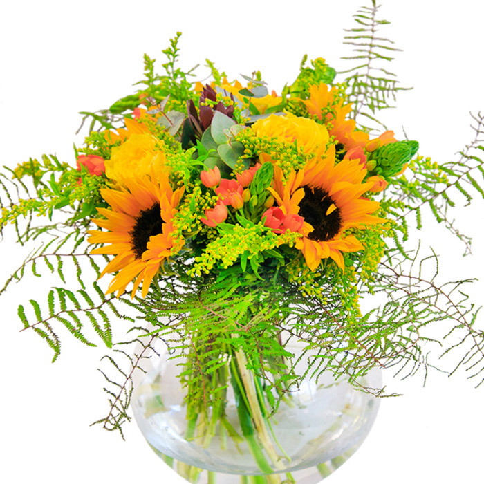 The initial impression of this arrangement is unmistakable-a sunburst of energy crafted by Yiewsley Florist, ideal for brightening tables from Horton Road to the banks of the canal. Sunflowers, their yellow-gold petals unfurled in generous layers, serve as the bouquet's joyful anchor, each bloom beaming with a distinct warmth. Beside them, garden roses in shades of yellow cradle their perfectly spiral forms, adding plush texture and a whisper of classic elegance. Scarlet red clusters of Hypericum berries nestle between the blooms, their smooth surfaces contrasting against soft petals and feathery green ferns that spill gracefully outward. Light green filler and stakes of solidago punctuate the composition with minute yellow stars, while bluish eucalyptus leaves lend a subtle woodland freshness, echoing the wild charm of local Yiewsley gardens. The crystal-clear, fishbowl vase unveils the architecture of the stems and the fresh, lively water below. If one could lean closer, the interplay of texture would be felt-the plushness of sunflower faces, the cushioned roses, the snap of berry stems, and the crisp greenness beneath. This sensory feast is delicately perfumed, conjuring memories of warm afternoons and celebratory gatherings. It is a perfect offering for birthdays, anniversaries, or any event where happiness should fill the room, brought to you from the flower-loving heart of Yiewsley.